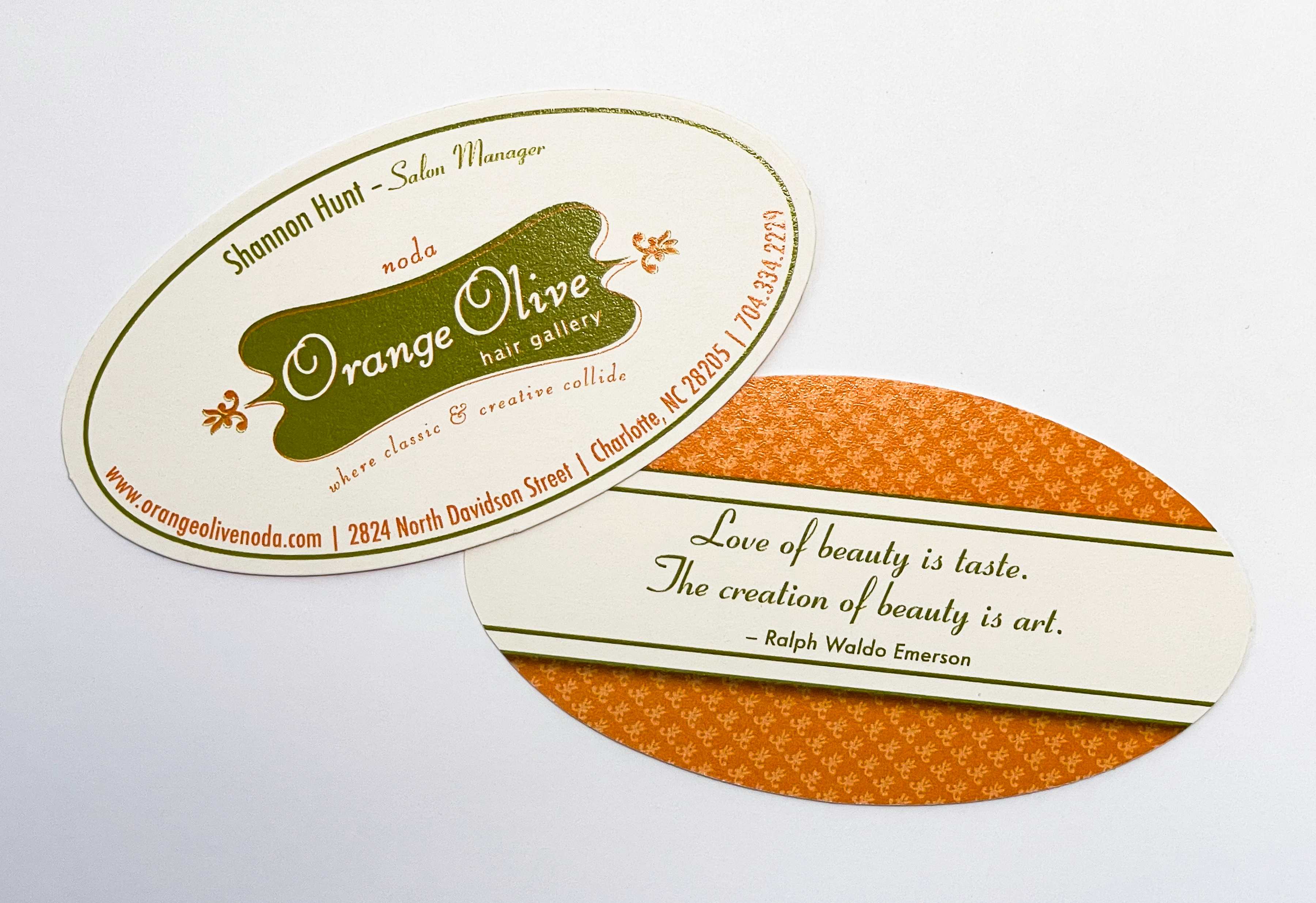 A narrow, oval-shaped business card featuring the logo for Orange Olive Hair Gallery on the front along with the name and title of the staff member and the contact information for the salon, all set in a ribbon-like typeface in bright orange and olive green. The back of the card features and orange fluer-de-lis pattern with a quote by Emerson that reads 'Love of beauty is taste. The creation of beauty is art.'